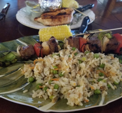A plate with a colorful kebab, rice with peas and carrots, and a corn on the cob, with grilled chicken in the background.