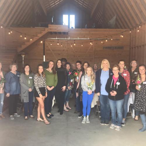 A group of people smiling and posing together inside a barn, decorated with string lights and holding flowers.