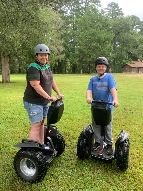 Two people, a woman and a boy, wearing helmets, stand on electric scooters in a grassy area with trees in the background.