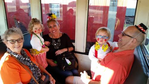 A group of four people, including two children in clown costumes, smiling and sitting together in a train car.