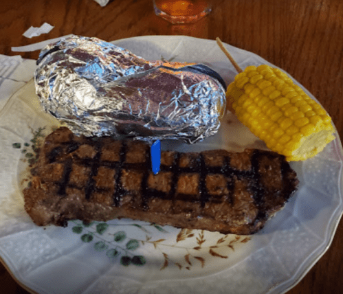 A plate with a grilled steak, a foil-wrapped baked potato, and a piece of corn on the cob.