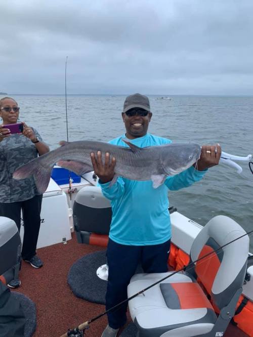 A man on a boat proudly holds a large catfish, while a woman takes a photo in the background.