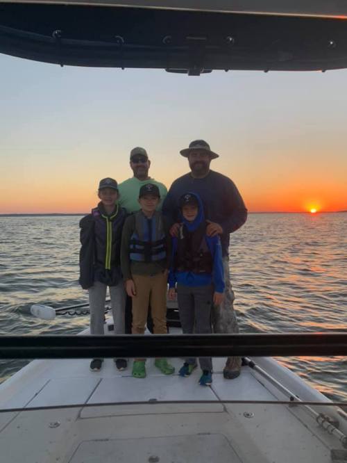 A group of four people, including two boys, stand on a boat at sunset, with water and a colorful sky in the background.
