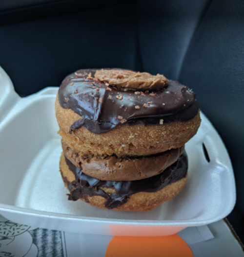 A stacked chocolate donut with a creamy filling, topped with chocolate icing and a cookie on top, in a takeout container.