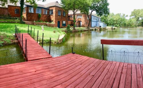 A red wooden dock extends over a calm pond, with rustic buildings and greenery in the background.
