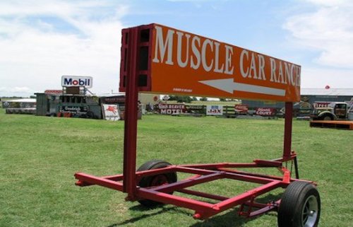 Sign for "Muscle Car Ranch" on a grassy field, with an arrow pointing to the right. Background features various buildings.
