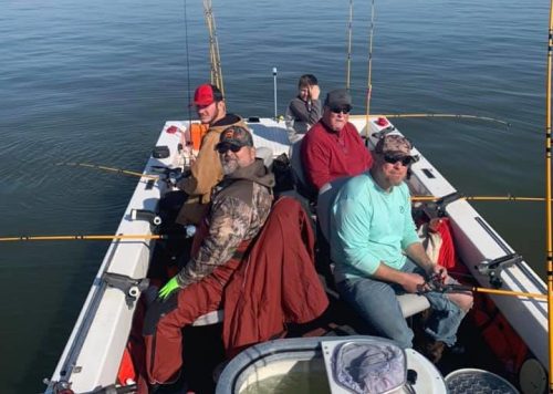 A group of five fishermen sitting in a boat on calm water, with fishing rods and gear around them under a clear blue sky.