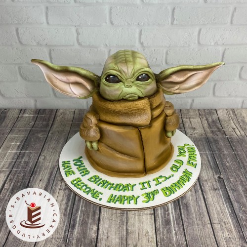 A birthday cake shaped like a character with large ears and green skin, wearing a brown robe, with a festive message.