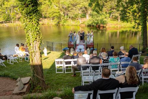 A wedding ceremony by a serene lake, with guests seated and a couple exchanging vows on a small platform.