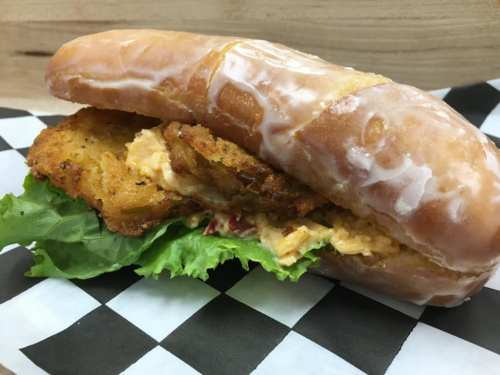 A glazed donut sandwich filled with fried chicken, lettuce, and a creamy spread, served on a checkered paper.