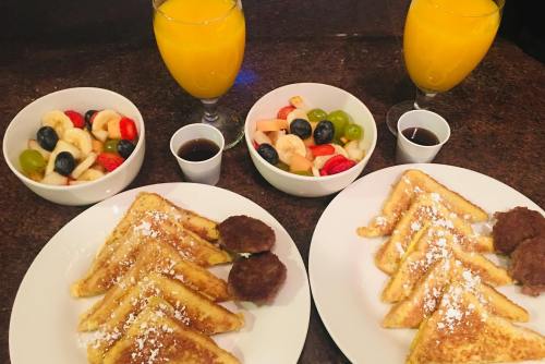 Two plates of French toast with powdered sugar, sausage patties, fruit bowls, and glasses of orange juice.