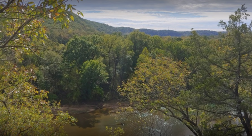 A scenic view of a river surrounded by lush green trees and rolling hills under a cloudy sky.