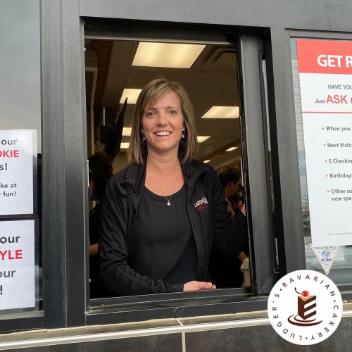 A smiling woman at a drive-thru window, wearing a black jacket, with promotional signs in the background.