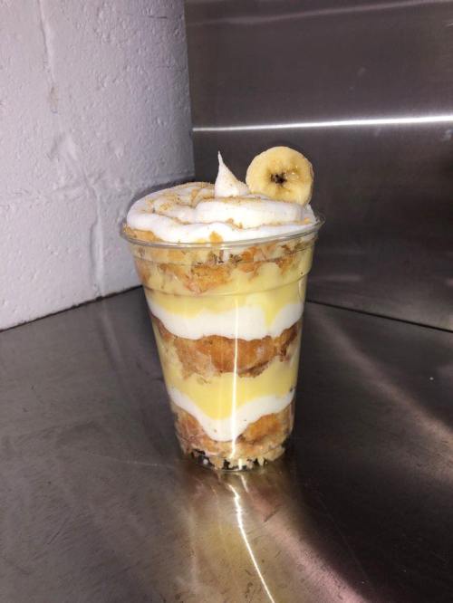 A layered dessert in a clear cup, featuring banana, cream, and crumbled cookies, topped with a banana slice.