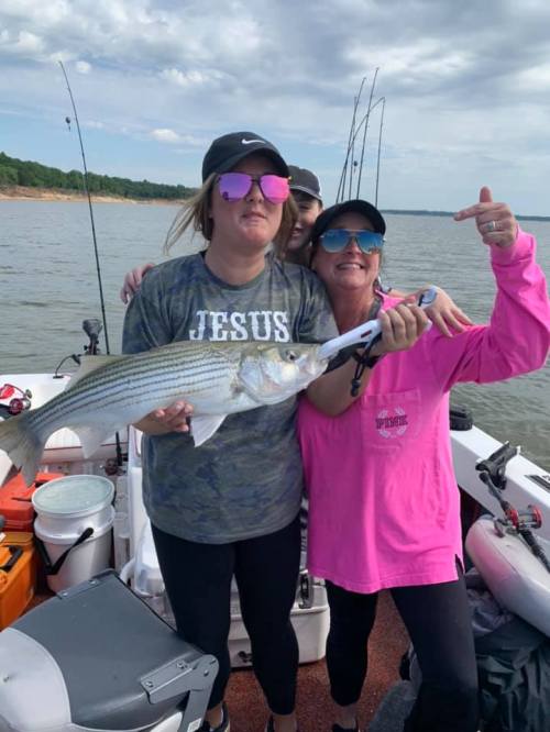 Three women on a boat hold a large fish, smiling and posing for the camera with fishing gear in the background.
