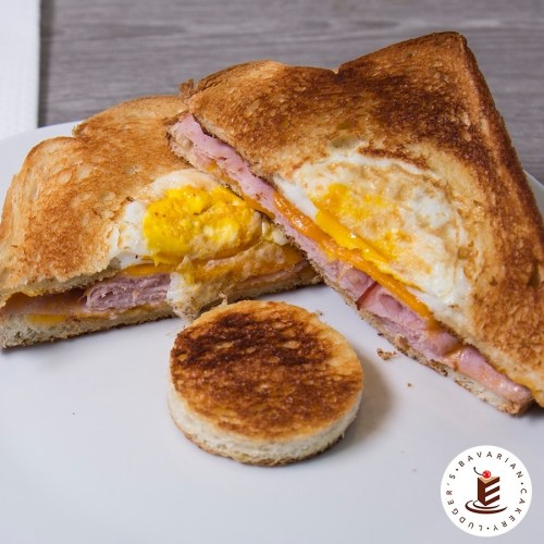 A toasted sandwich filled with ham, cheese, and a fried egg, served with a small round piece of toasted bread.
