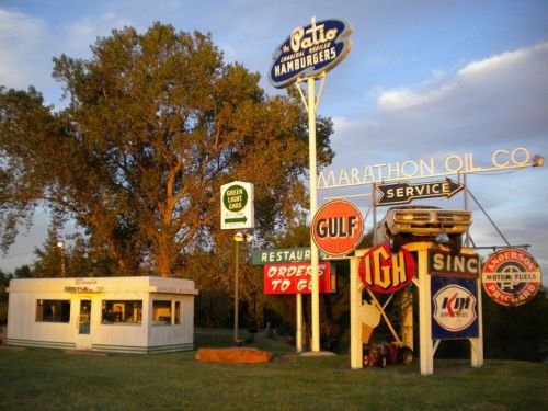 Vintage roadside scene featuring a hamburger stand and various gas station signs at sunset.