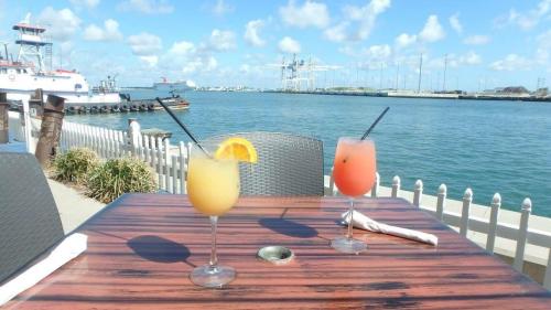Two colorful cocktails on a wooden table by the water, with boats and a clear blue sky in the background.
