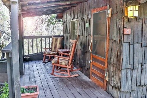 A rustic wooden porch with two rocking chairs and a door, surrounded by trees and soft sunlight.