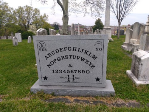 A gray gravestone with letters and numbers, featuring "YES" and "NO" symbols, set in a grassy cemetery.