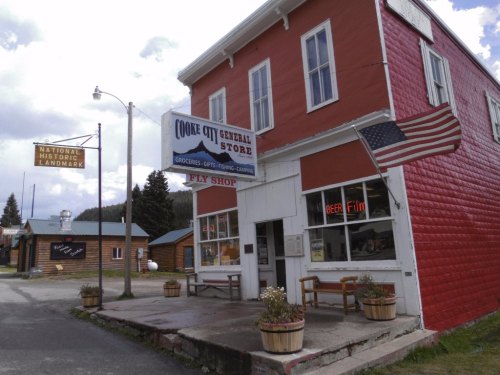 A rustic general store with a red exterior, featuring a sign for groceries and a flag, set in a small town.