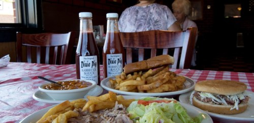 A table with barbecue dishes: pulled pork, fries, a sandwich, baked beans, and two bottles of barbecue sauce.