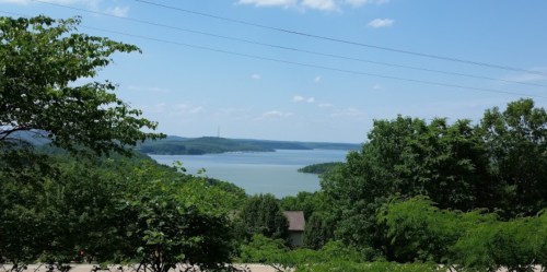 A scenic view of a lake surrounded by green trees and hills under a clear blue sky with a few clouds.