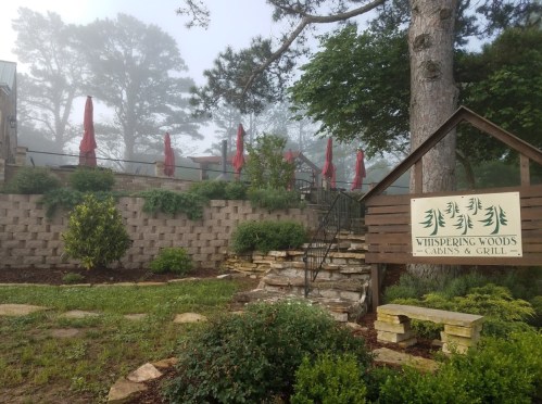 A foggy scene of Whispering Woods Cabins & Grill, featuring a sign, stone steps, and red umbrellas on a patio.