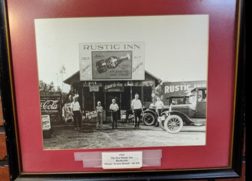 Black and white photo of five men outside the Rustic Inn, with vintage cars and signage from the early 1900s.
