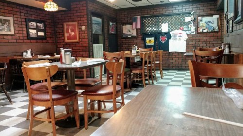 A cozy restaurant interior with wooden tables and chairs, brick walls, and checkered flooring, featuring framed photos.