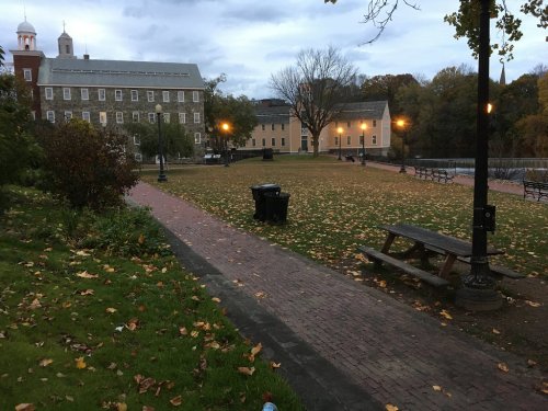 A park with benches and a trash can, surrounded by trees and buildings, under a cloudy sky with fallen leaves on the ground.