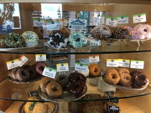 A display case filled with various colorful donuts, labeled with names like "Maple Fried" and "Chocolate Sprinkle."