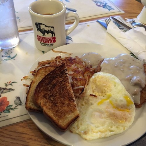 A plate of breakfast featuring toast, hash browns, a fried egg, and sausage gravy, with a coffee cup in the background.