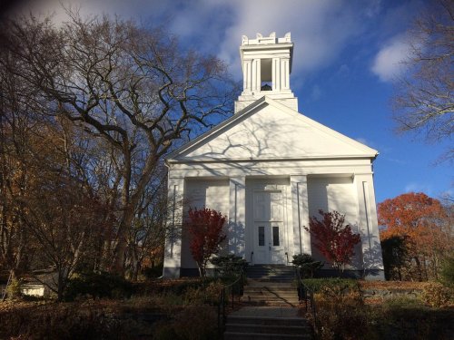 A white church building with a bell tower, surrounded by trees and autumn foliage under a blue sky.