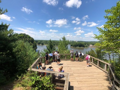 A scenic overlook with people enjoying the view of a river and lush greenery under a blue sky with fluffy clouds.