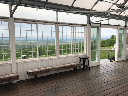 A spacious indoor area with large windows overlooking a green landscape and cloudy sky. Wooden benches and a stool are visible.