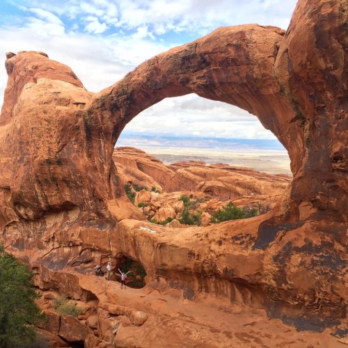 A natural rock arch in a desert landscape, with two small figures standing beneath it against a cloudy sky.