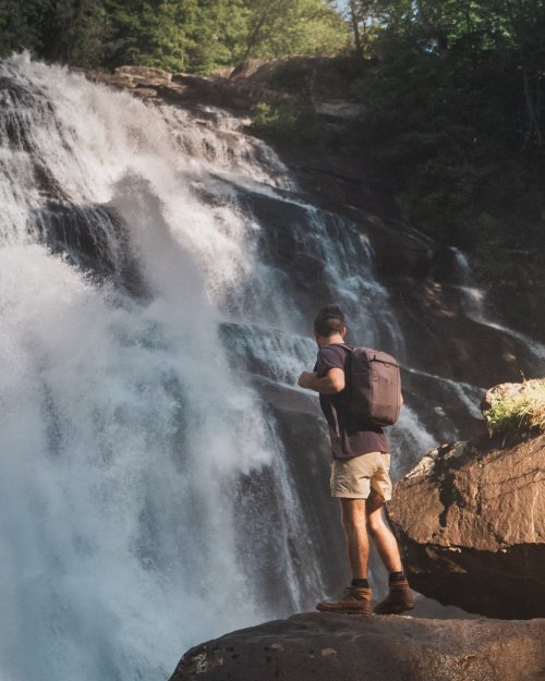 A person stands on a rock, gazing at a large waterfall surrounded by lush greenery.
