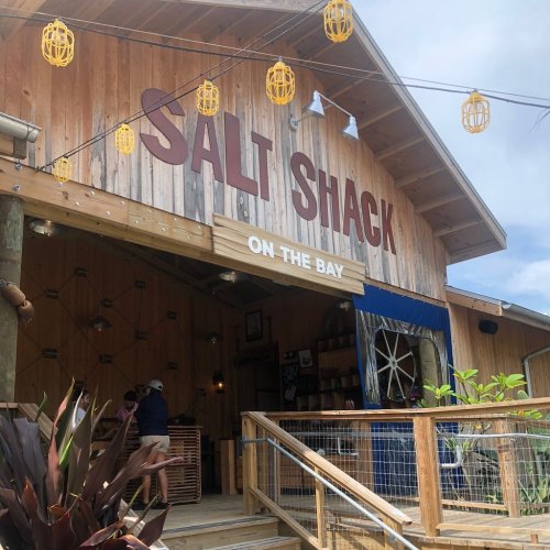 A wooden building with "SALT SHACK ON THE BAY" sign, featuring outdoor seating and decorative lights.