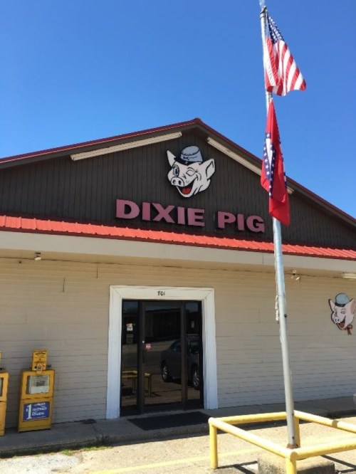 Exterior of Dixie Pig restaurant with a pig mascot sign, American and Arkansas flags, and a clear blue sky.
