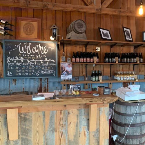 A rustic bar with a chalkboard welcome sign, shelves of bottles, and wooden decor in a cozy tasting room.