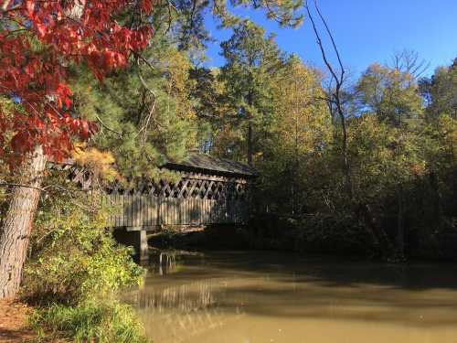 A wooden covered bridge spans a calm river, surrounded by trees with autumn foliage under a clear blue sky.