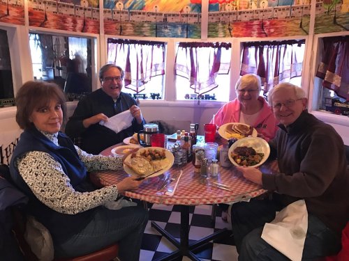 Four people sitting at a round table in a diner, smiling and holding plates of food. Colorful decor in the background.