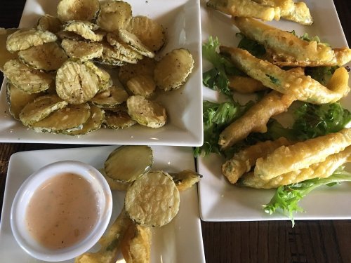 Three plates of fried vegetables: crispy zucchini chips, fried pickles with dipping sauce, and battered green beans on lettuce.