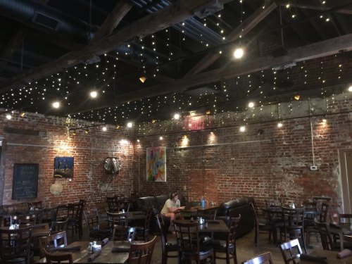 Cozy restaurant interior with exposed brick walls, string lights, and wooden tables. A person sits at a table.