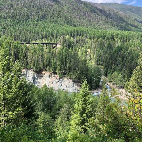 A lush green landscape with a river and a bridge nestled among mountains and dense trees under a clear blue sky.