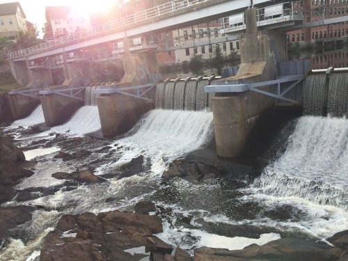 A dam with multiple spillways releasing water, surrounded by rocky terrain and buildings in the background.