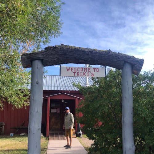 A person stands under a wooden archway with a sign reading "Welcome to Trix's," in front of a red building.