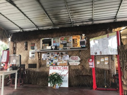 A rustic food stand with a thatched roof, featuring a menu board and various flyers on the walls.