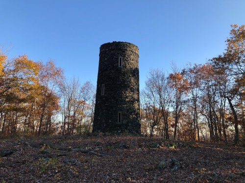 A tall, stone tower stands amidst autumn trees with colorful leaves under a clear blue sky.